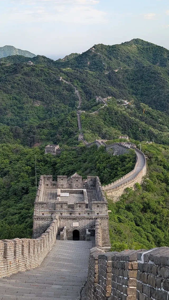 The Great Wall of China at Mutianyu snaking over green forested mountain ridges into the distance, with a stone watchtower in the foreground, the kind of place that sparks an endless stream of on-demand history questions